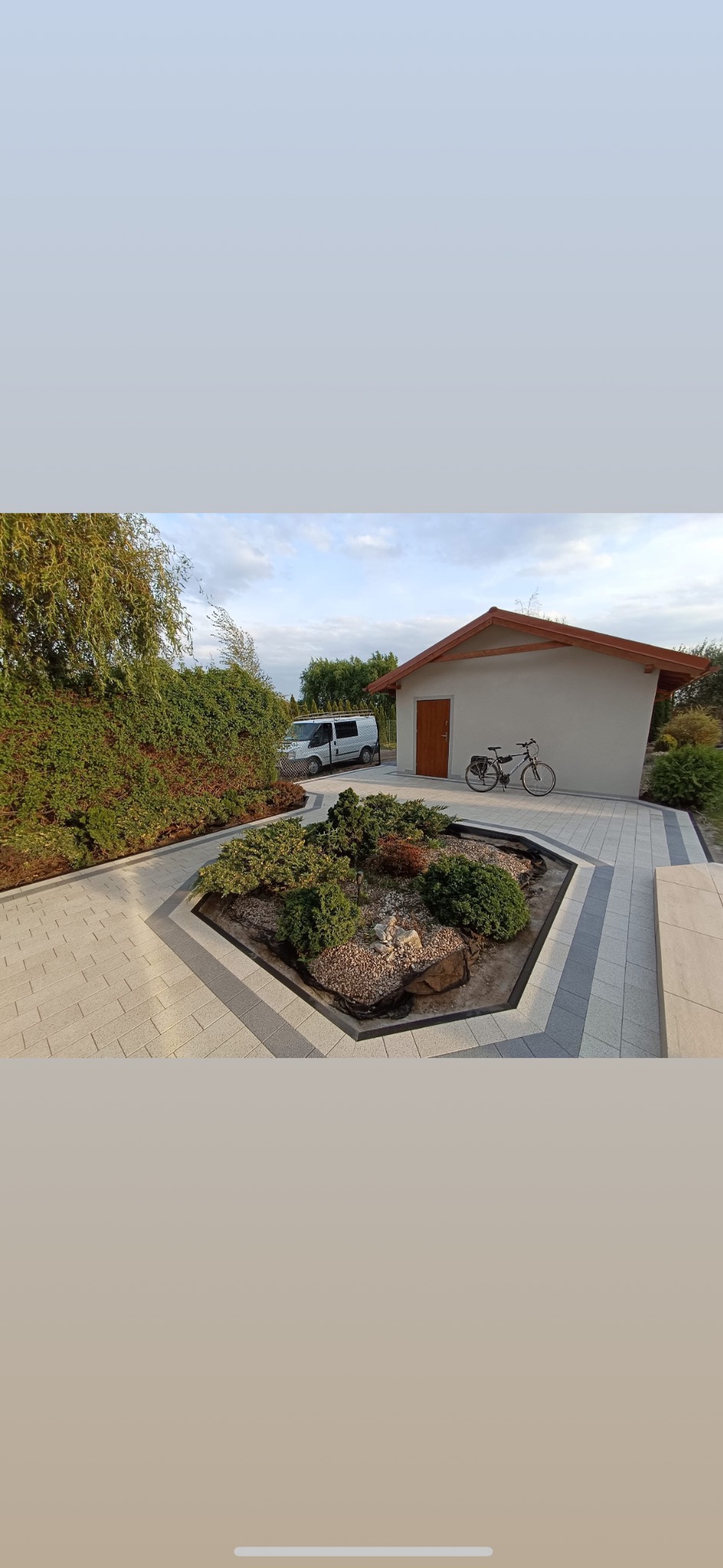 Jasna kostka brukowa otacza rabatę z kamieniami i zielenią. W tle budynek z rowerem i auto dostawcze. Kadr z perspektywy lekko z góry.