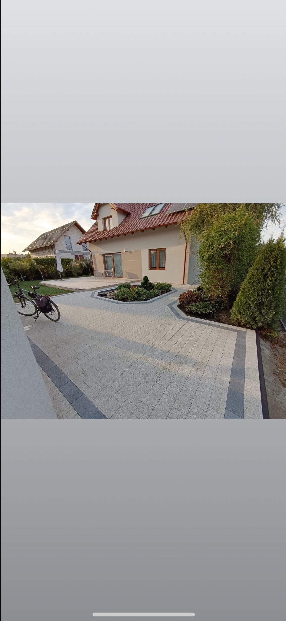 Jasnoszara kostka brukowa ułożona na podjeździe do domu z rabatą kwiatową pośrodku. Ciemniejsze obrzeża kostki. W tle dom z czerwoną dachówką i rower oparty o ścianę.