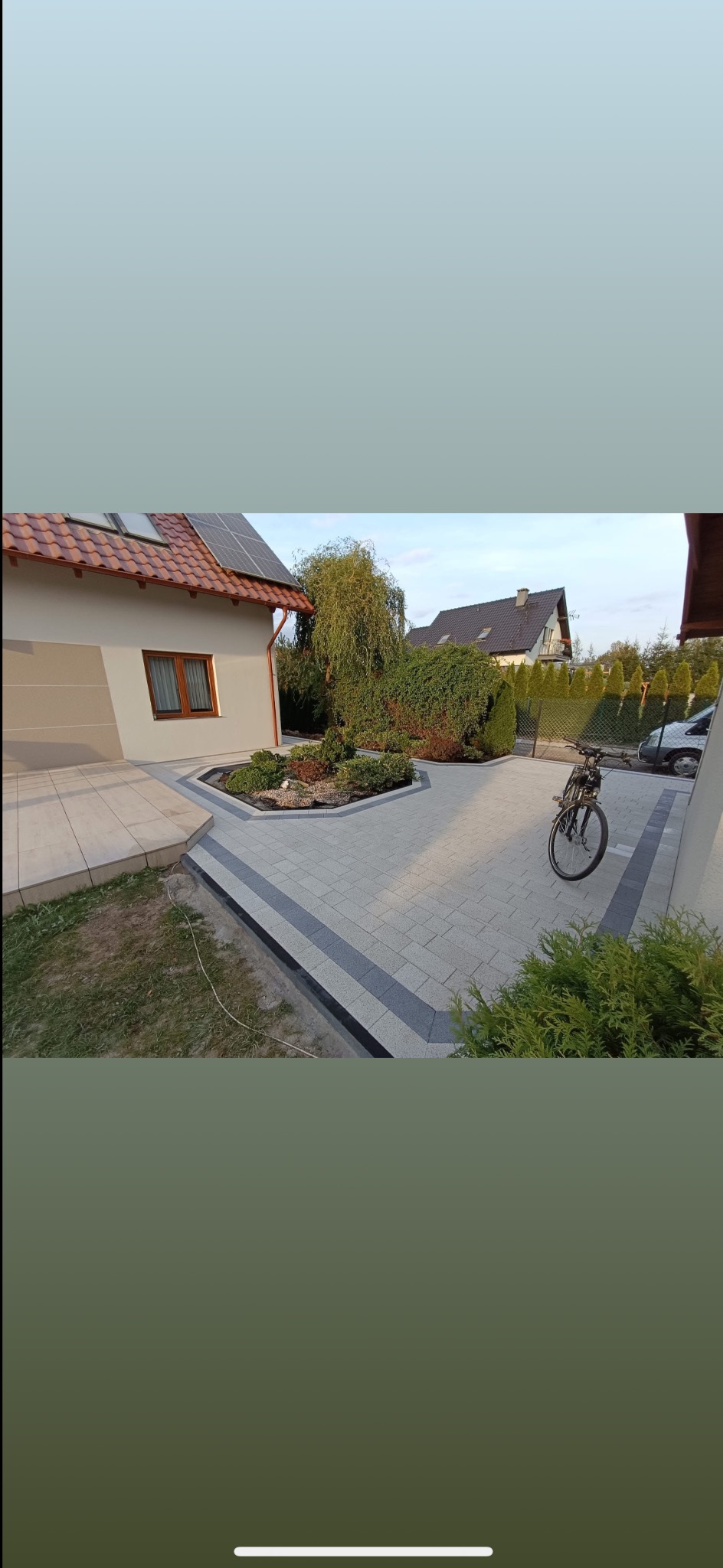 Jasnoszara kostka brukowa z ciemniejszym pasem, tworząca ścieżkę wokół rabaty z roślinami. W tle dom z panelami słonecznymi i rower.
