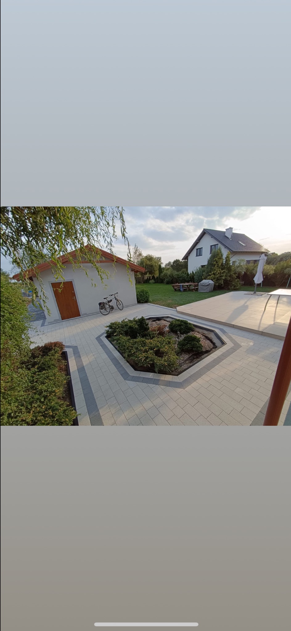 Jasna kostka brukowa ułożona w geometryczny wzór wokół rabaty z krzewami. W tle budynek gospodarczy i dom jednorodzinny.