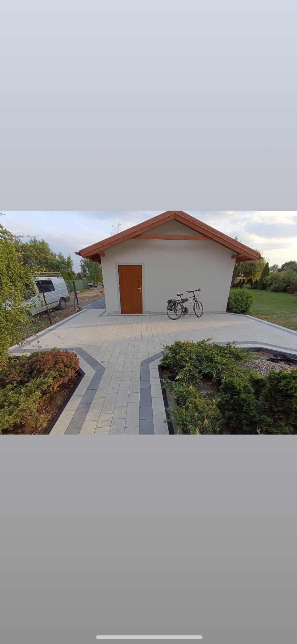 Ułożona kostka brukowa w Podjazach, tworząca ścieżkę do budynku z drewnianymi drzwiami. Widoczny rower oparty o ścianę i starannie zaplanowana zieleń.