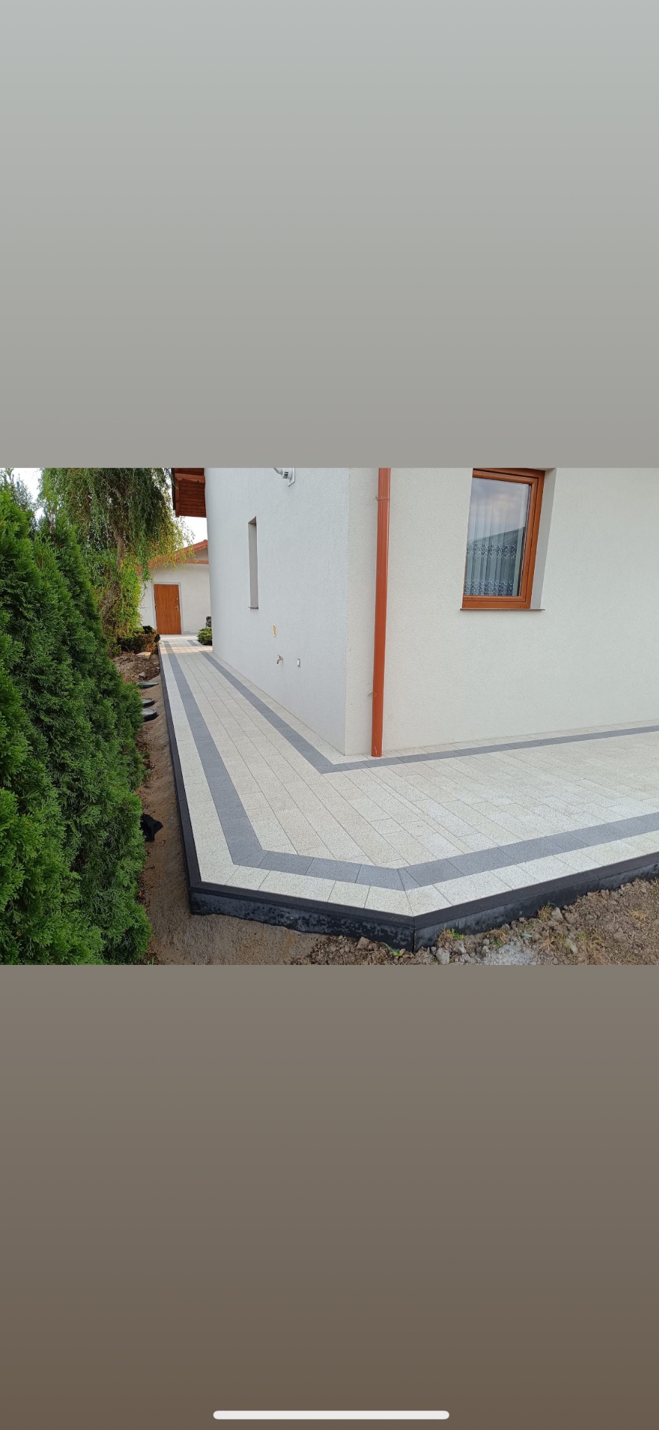 Jasna kostka brukowa z szarym obramowaniem okalająca dom, widok z boku. Z prawej strony brązowa rynna, z lewej tuje.