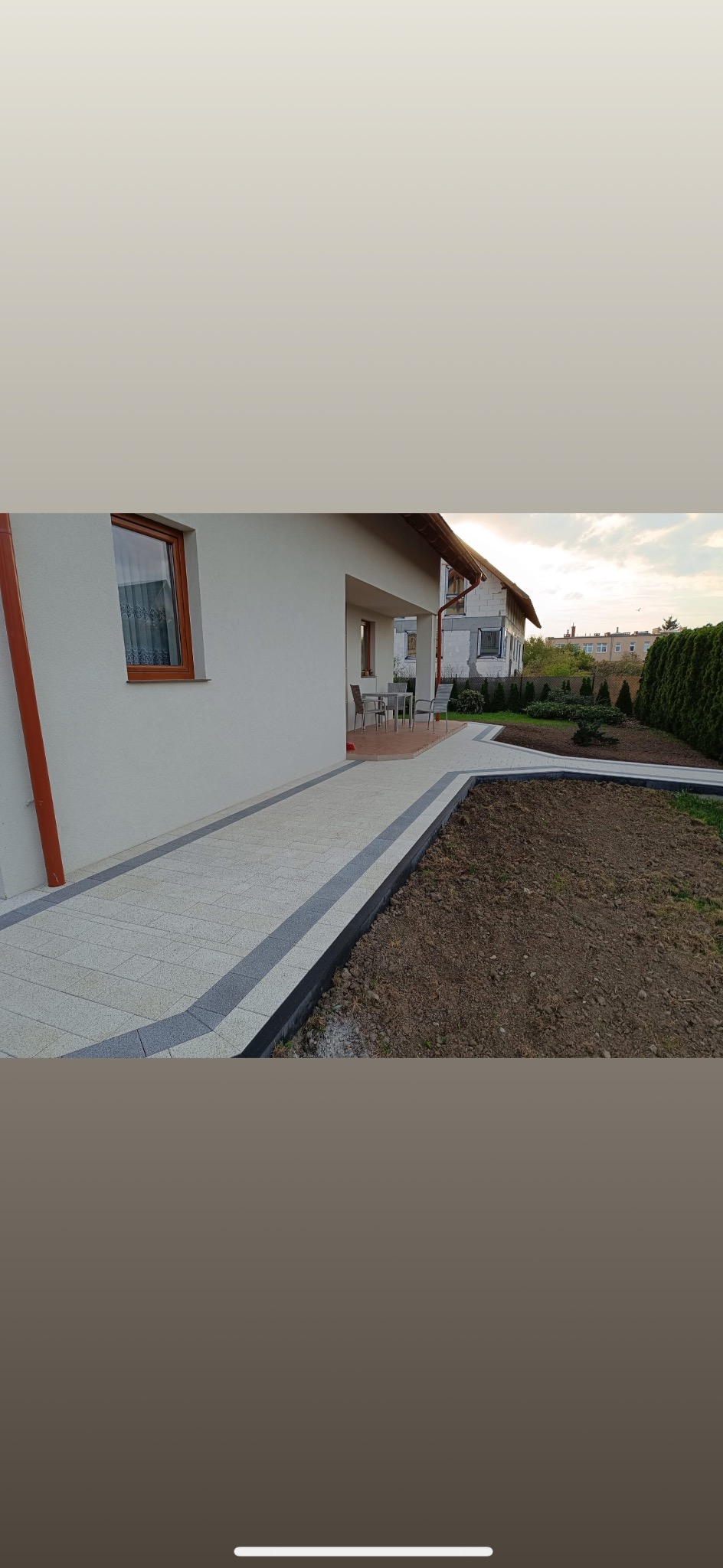 Jasna kostka brukowa z ciemnoszarym obramowaniem prowadząca do domu. Przygotowany teren pod nasadzenia. Widok na budynek i fragment ogrodu.