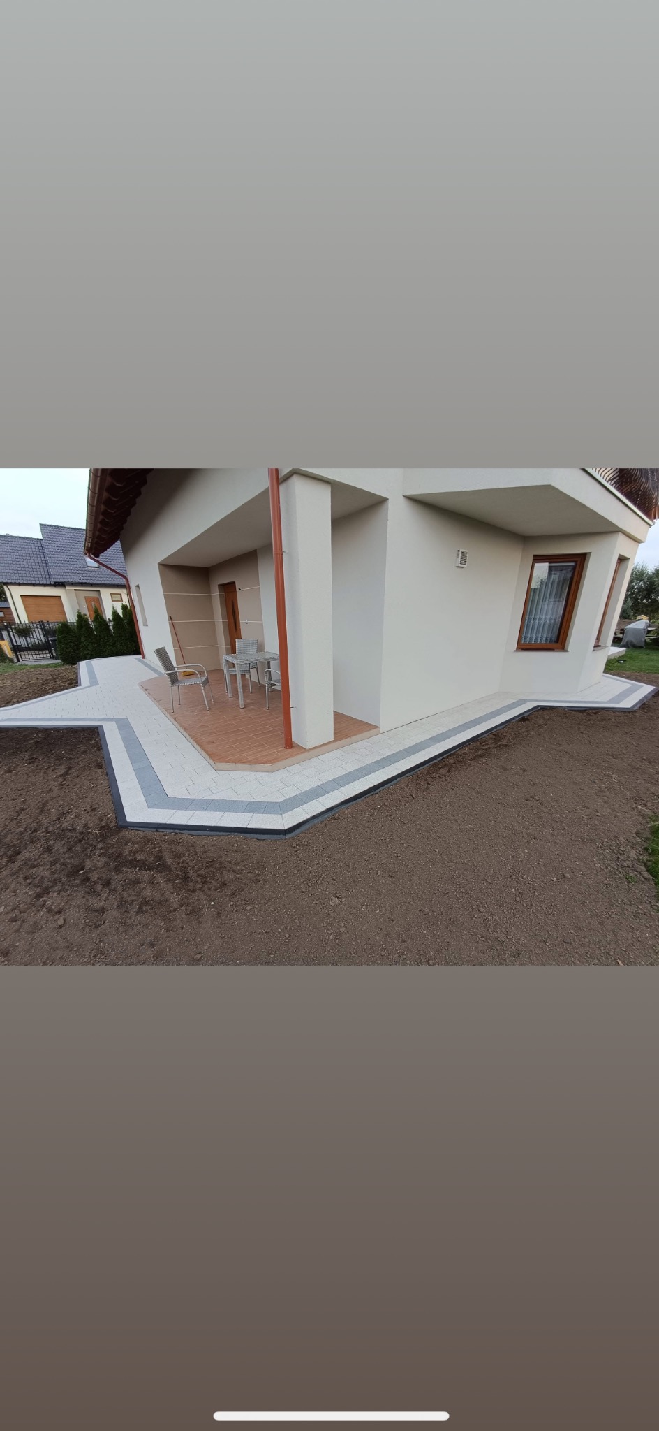 Wykończony taras z kostki brukowej w odcieniach szarości i beżu, przylegający do jasnego budynku z brązowymi elementami. Widoczne meble ogrodowe i świeżo przygotowana ziemia.