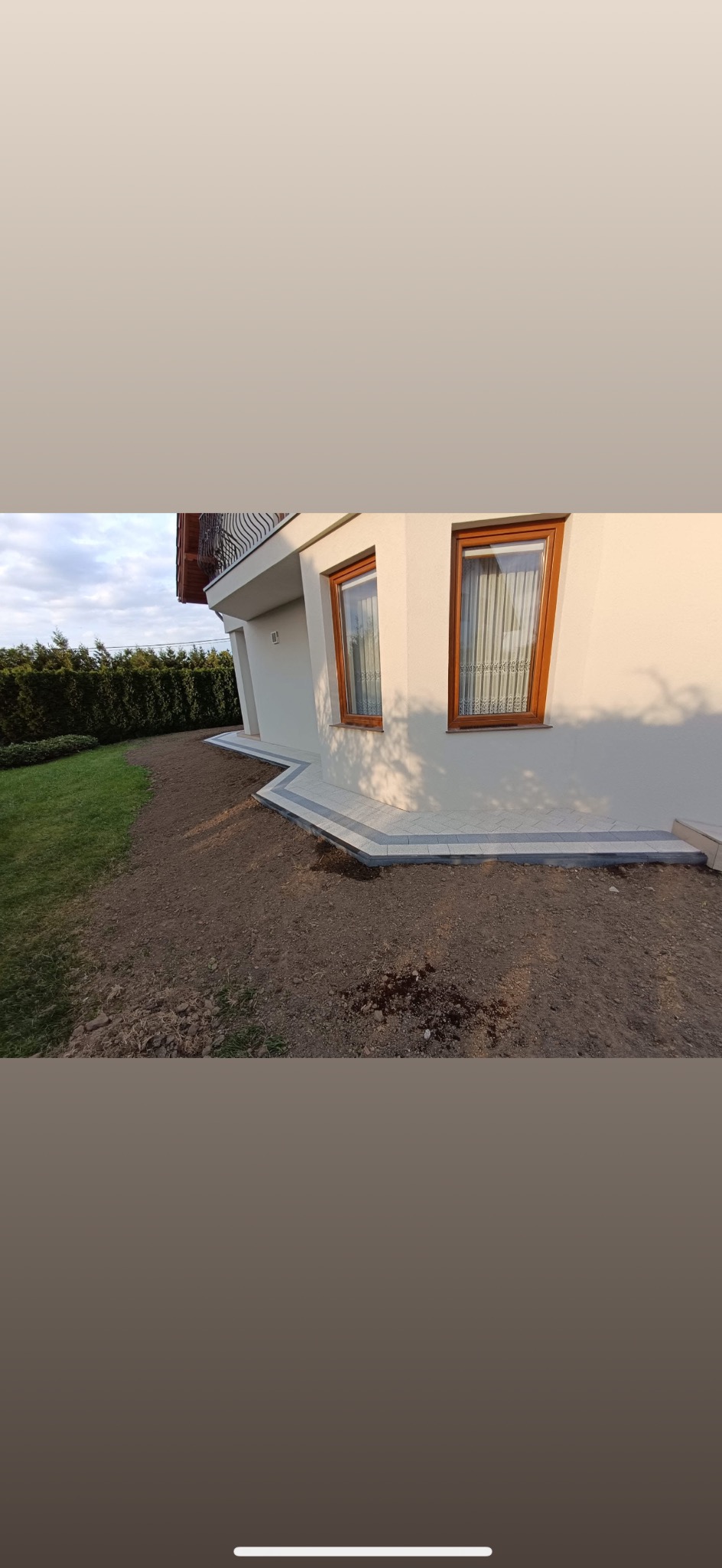 Jasna elewacja domu z dwoma oknami i obłożonym kostką brukową obrzeżem. Kostka w odcieniach szarości, trawnik i rabata w tle.