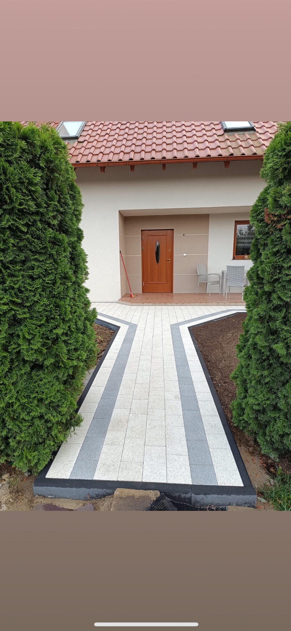 Ścieżka z jasnej kostki brukowej z szarymi pasami prowadząca do brązowych drzwi wejściowych domu z czerwoną dachówką, otoczona zielonymi tujami.