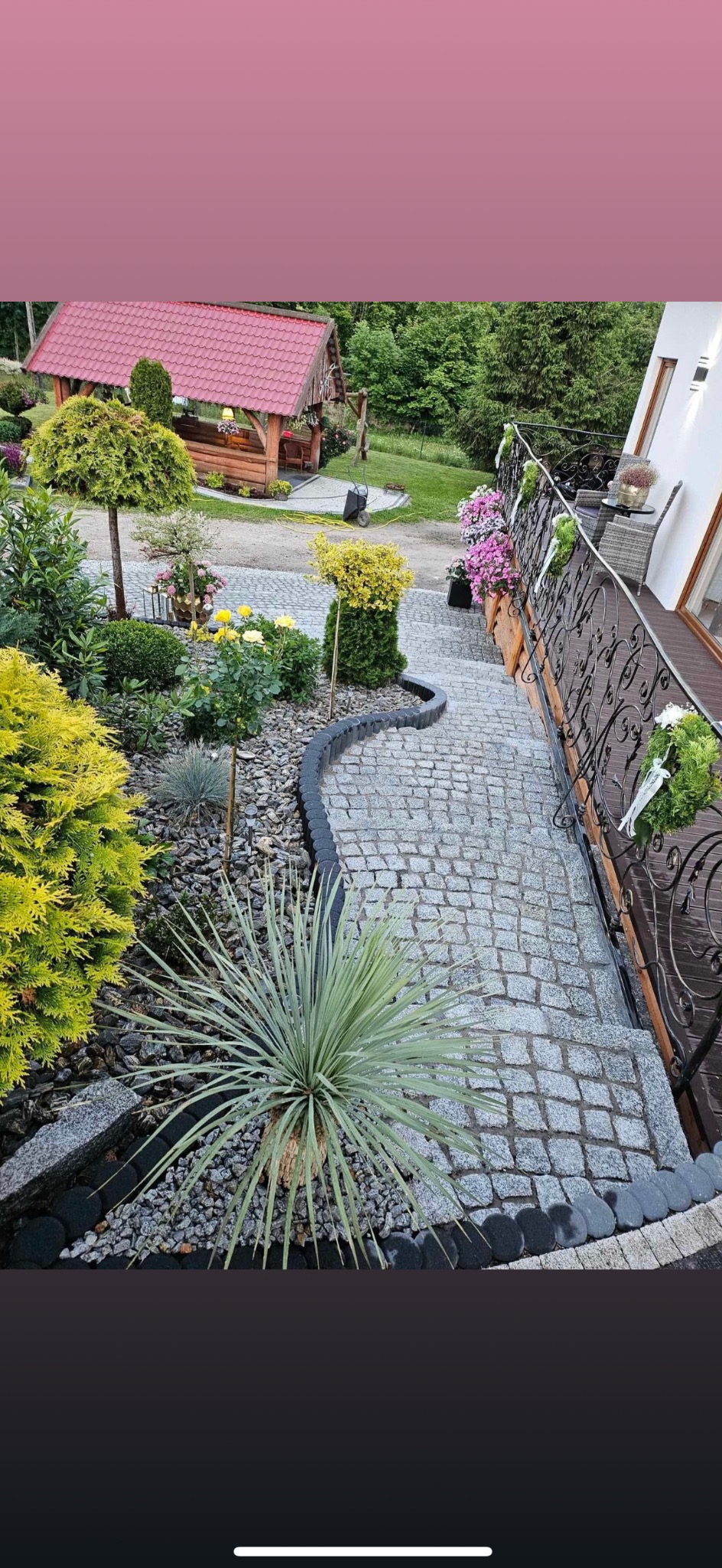 Ścieżka z kostki granitowej z ozdobnymi obrzeżami, prowadząca schodami w dół w zadbanym ogrodzie z altaną w tle.