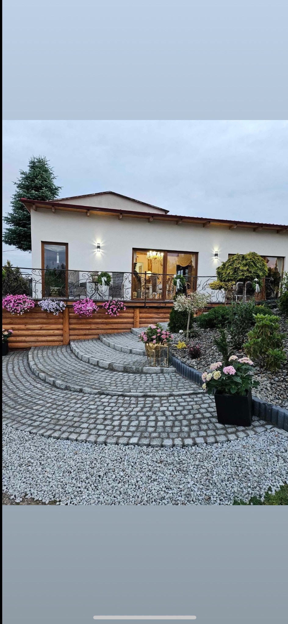 Elegancki podjazd z kostki granitowej w kształcie wachlarza prowadzący do domu. Widoczne schody z kostki, ozdobne rośliny i elementy drewnianego ogrodzenia balkonu.