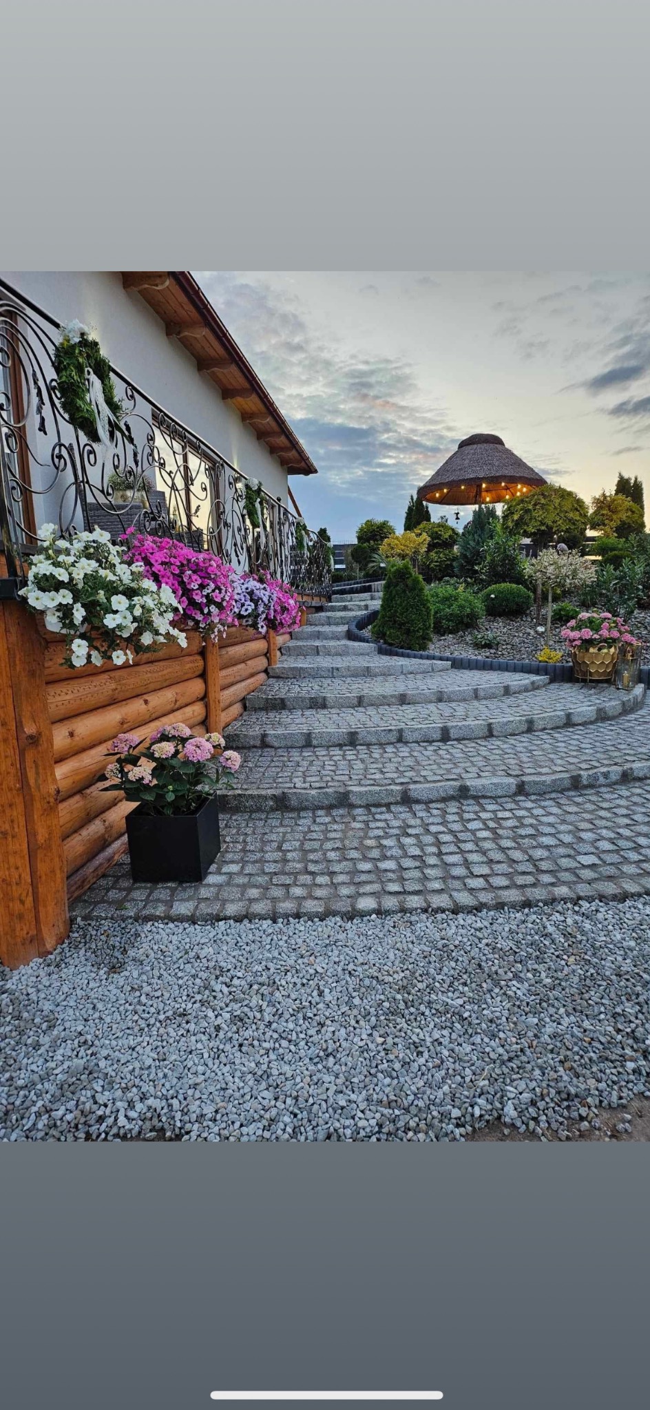 Granitowe schody prowadzą do altany w ogrodzie. Kwitnące kwiaty w donicach zdobią wejście i balustradę. Tło stanowi zadbana zieleń i niebo o zmierzchu.