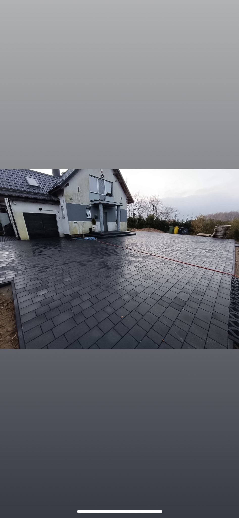 Ułożona, grafitowa kostka brukowa na podjeździe przed domem z jasną elewacją. Widoczne obrzeża i elementy drenażowe. Całość sprawia wrażenie solidnego wykończenia.