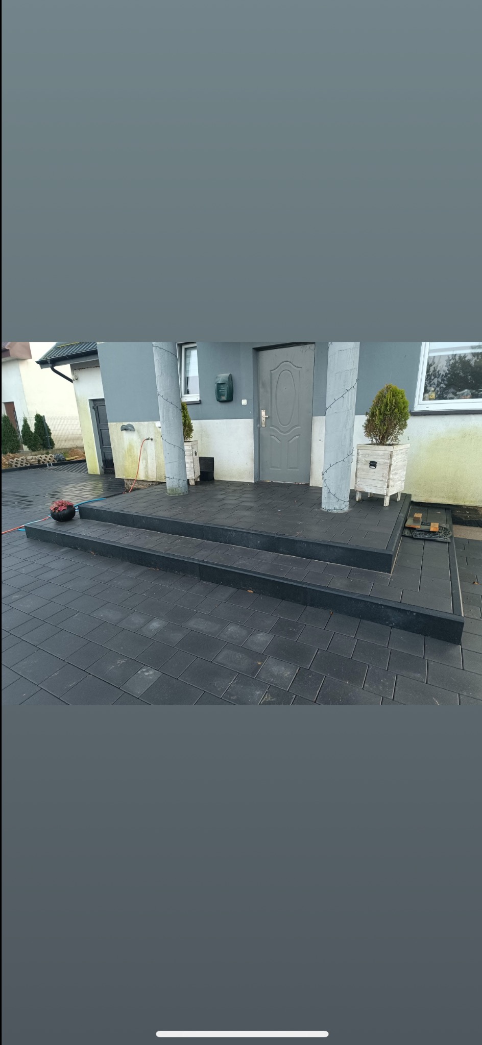 Ułożona, grafitowa kostka brukowa przed wejściem do domu, tworząca schody. Dwa betonowe słupy dekorowane lampkami. Donice z roślinami.