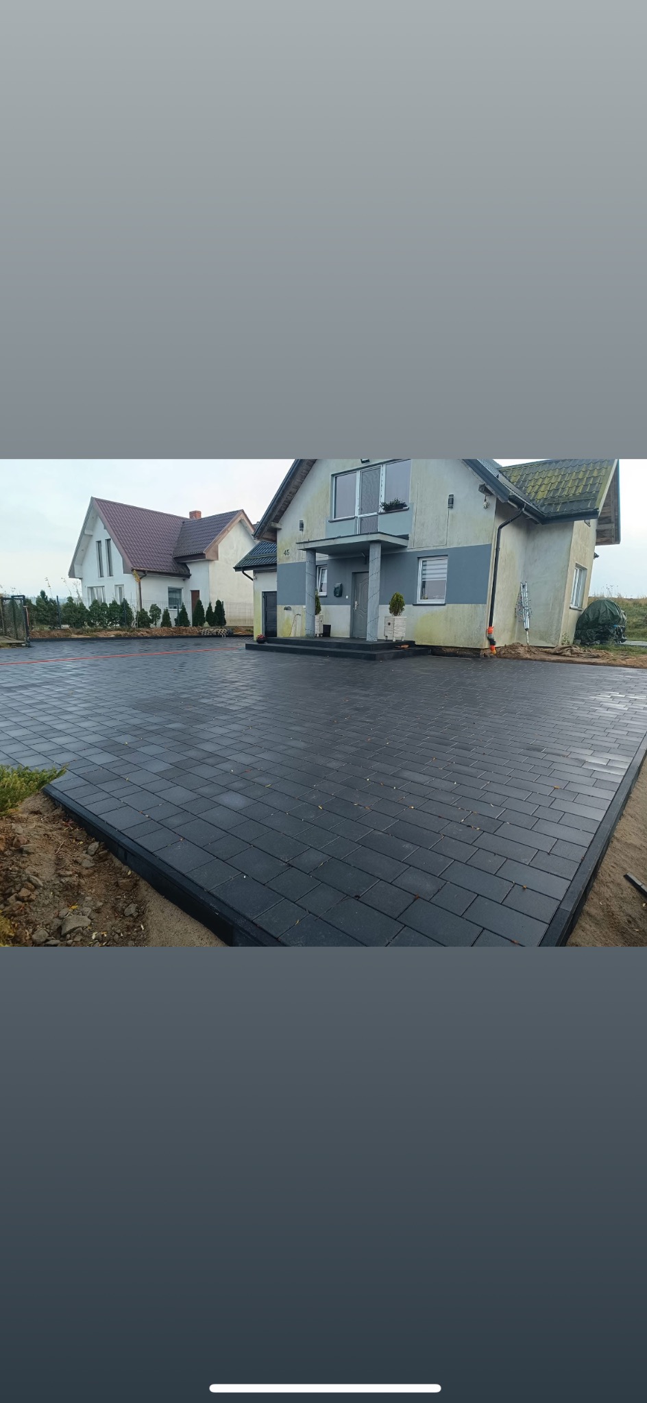 Ułożona, ciemnoszara kostka brukowa na podjeździe przed domem. Widoczne obrzeża i dwa budynki w tle. Ujęcie z poziomu gruntu.