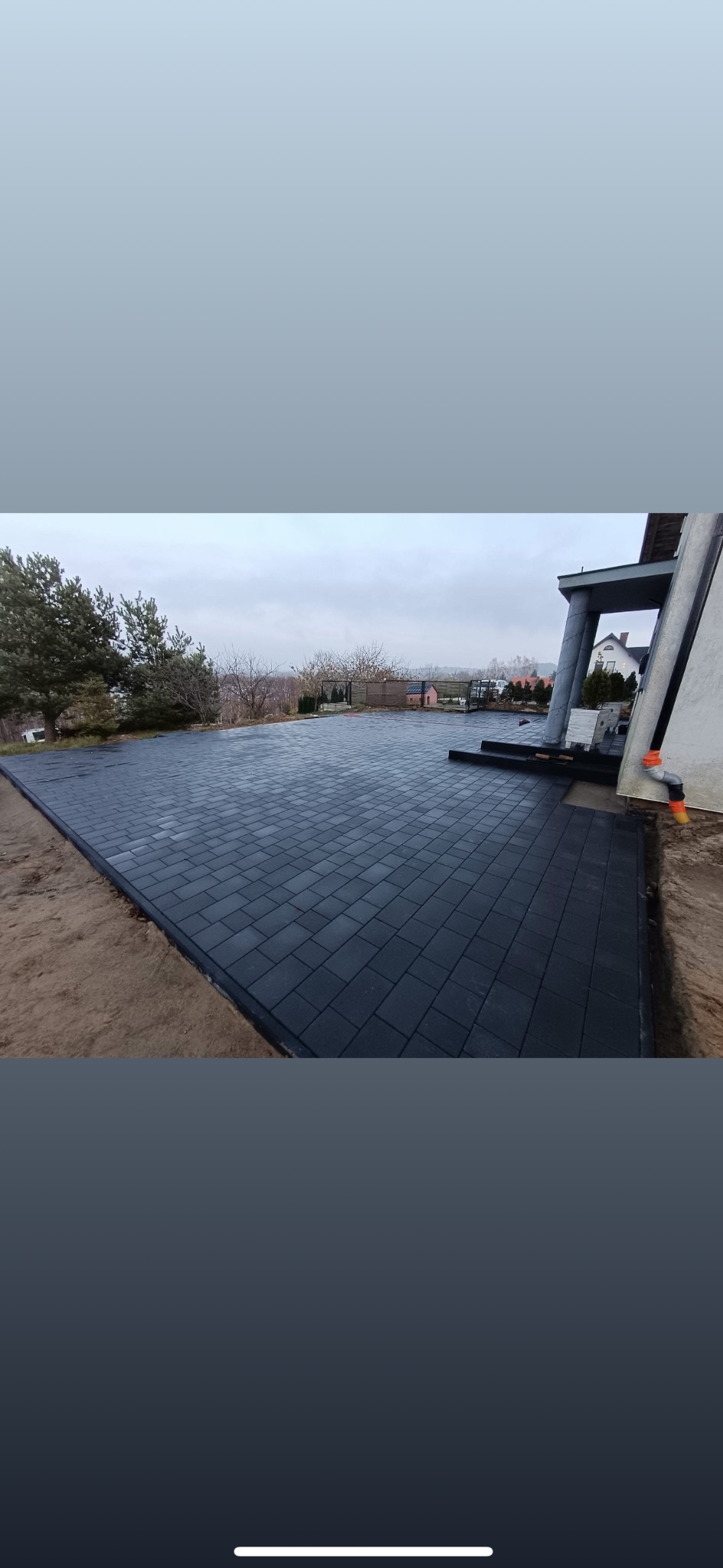 Rozległy taras z ciemnej kostki brukowej, obramowany piaskiem, przylegający do nowoczesnego domu z widokiem na zalesione wzgórze w tle.