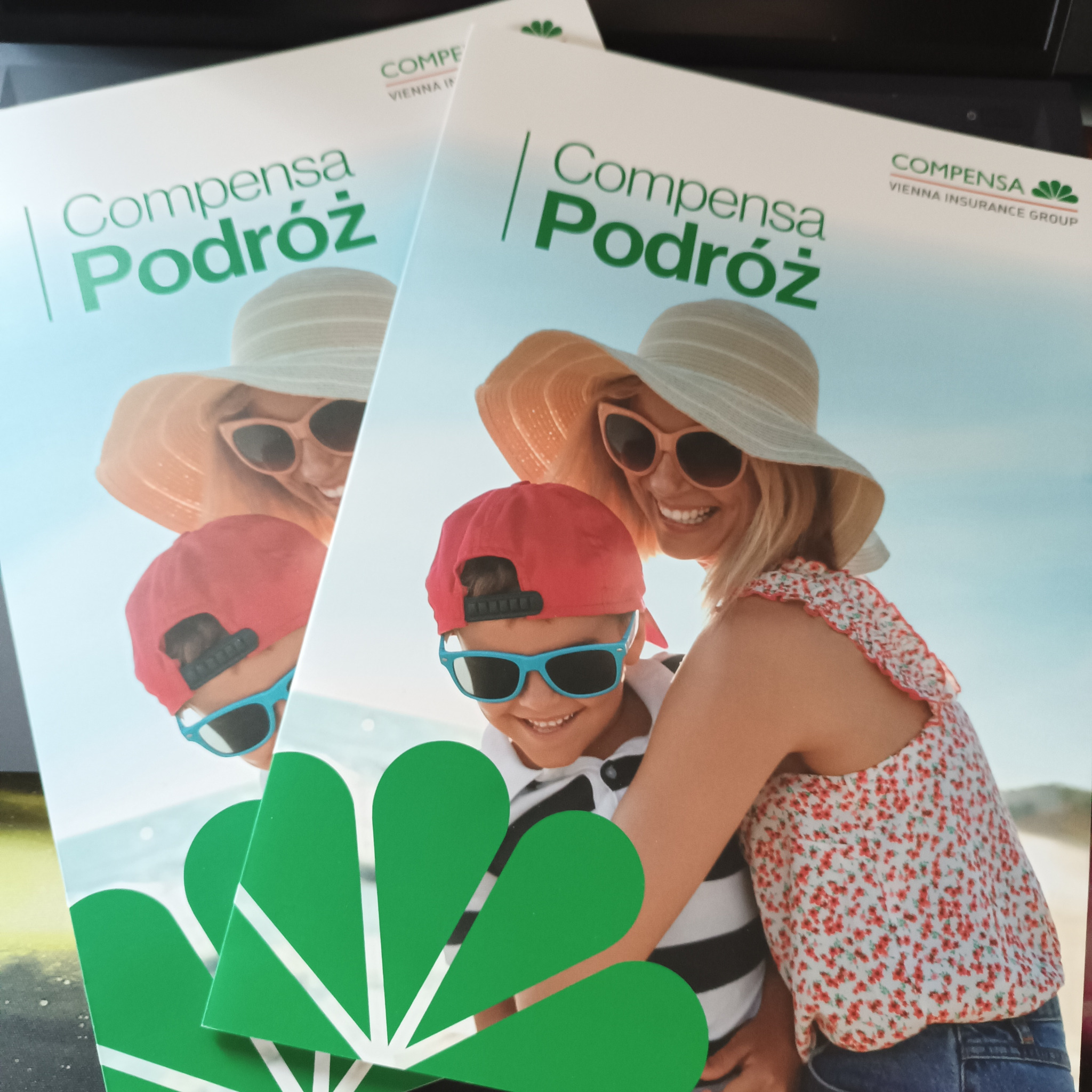 Ujęcie z góry na dwa foldery reklamowe firmy Compensa, promujące ubezpieczenia podróżne, z wizerunkiem uśmiechniętej matki w kapeluszu i syna w czapce i okularach przeciwsłonecznych na tle plaży.