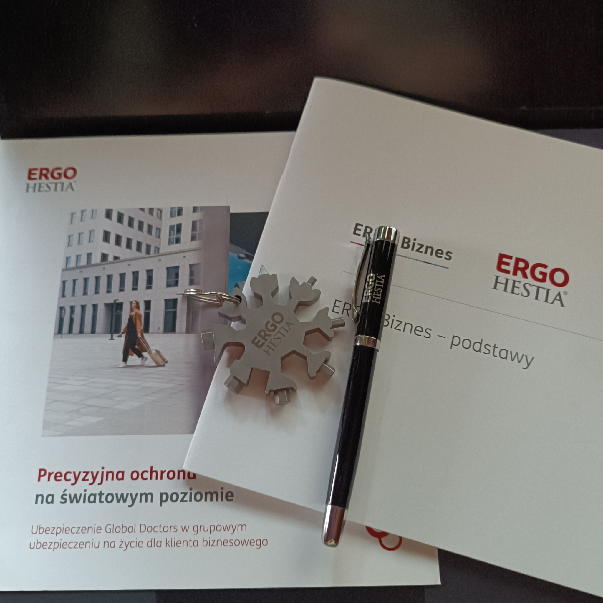 Ulotka informacyjna z logo Ergo Hestia leżąca obok breloczka w kształcie śnieżynki z logo firmy oraz czarnego długopisu.