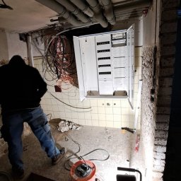 Tomasz Kapella Usługi wielobranżowe - Otwarta skrzynka elektryczna w trakcie montażu na kafelkowanej ścianie, widoczne kable, rury pod sufitem i narzędzia. Elektryk w tle.