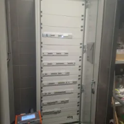 Otwarta szafa elektryczna z licznymi bezpiecznikami i miernikiem MPI-535 na wiadrach. Widoczne szare płytki na ścianie i regał z artykułami.