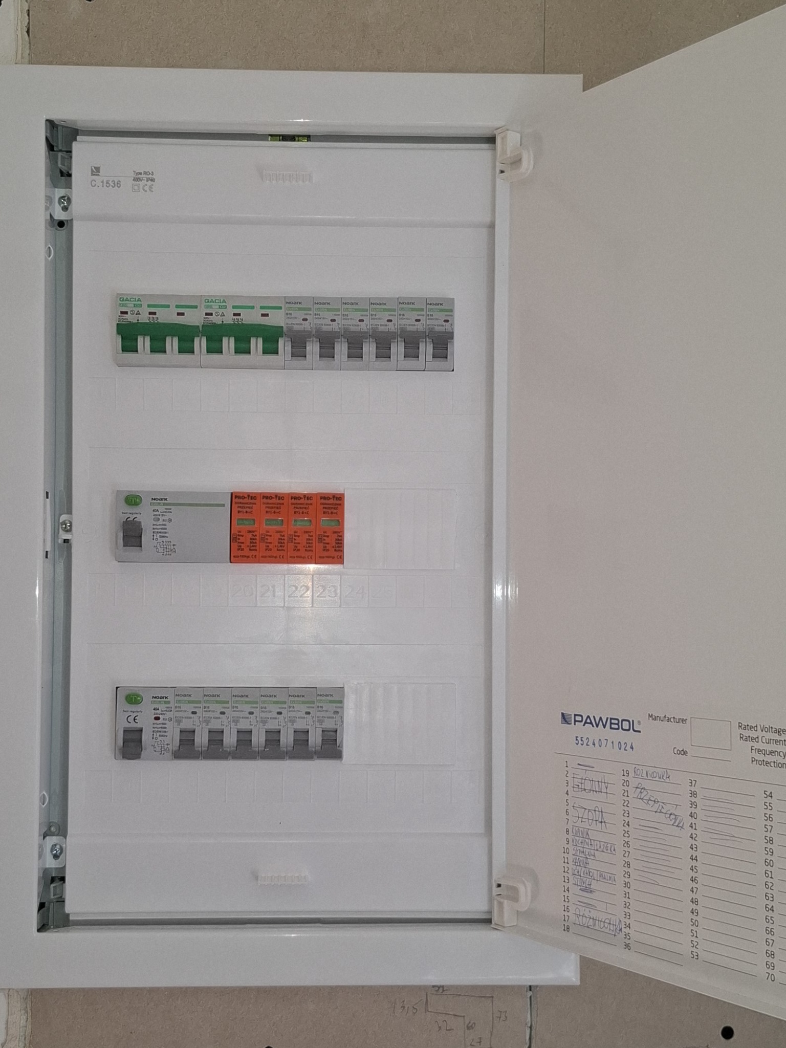 Otwarta skrzynka elektryczna z widocznymi bezpiecznikami i schematem, zamontowana na ścianie. Widoczne elementy instalacji elektrycznej.