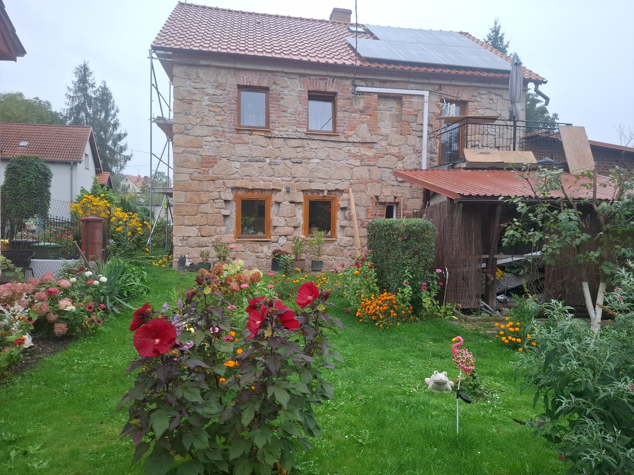 Kamienny dom z panelami słonecznymi na dachu w trakcie montażu. Widoczne rusztowania, kwiaty w ogrodzie i częściowo odnowiona elewacja.
