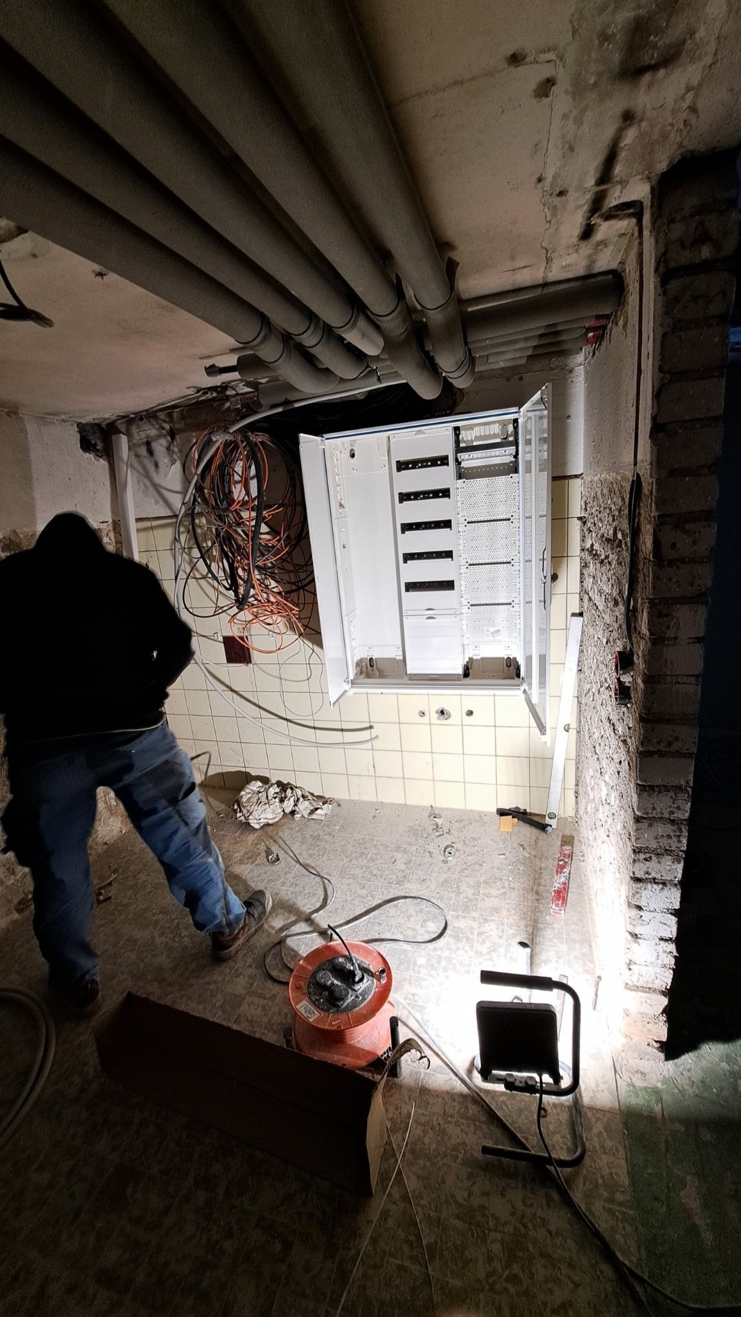 Otwarta skrzynka elektryczna w trakcie montażu na kafelkowanej ścianie, widoczne kable, rury pod sufitem i narzędzia. Elektryk w tle.