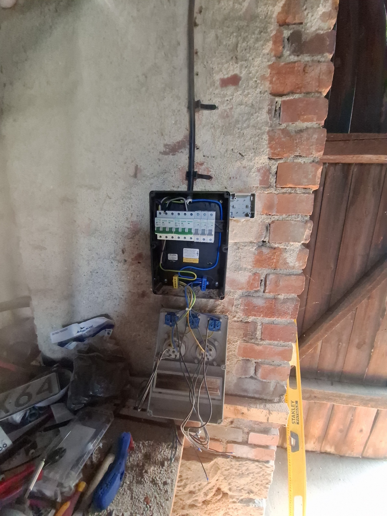 Otwarta skrzynka elektryczna z bezpiecznikami na ceglanej ścianie, widoczne przewody i narzędzia. Użycie poziomicy dla precyzji montażu instalacji elektrycznej.