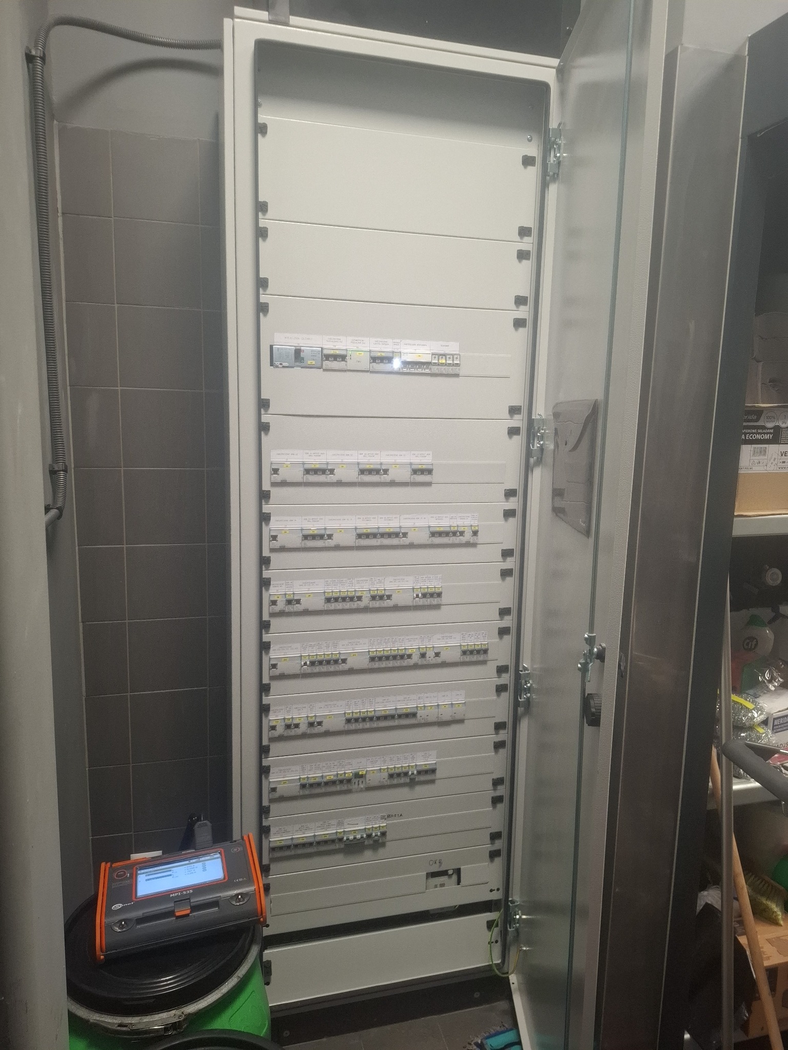 Otwarta szafa elektryczna z licznymi bezpiecznikami i miernikiem MPI-535 na wiadrach. Widoczne szare płytki na ścianie i regał z artykułami.