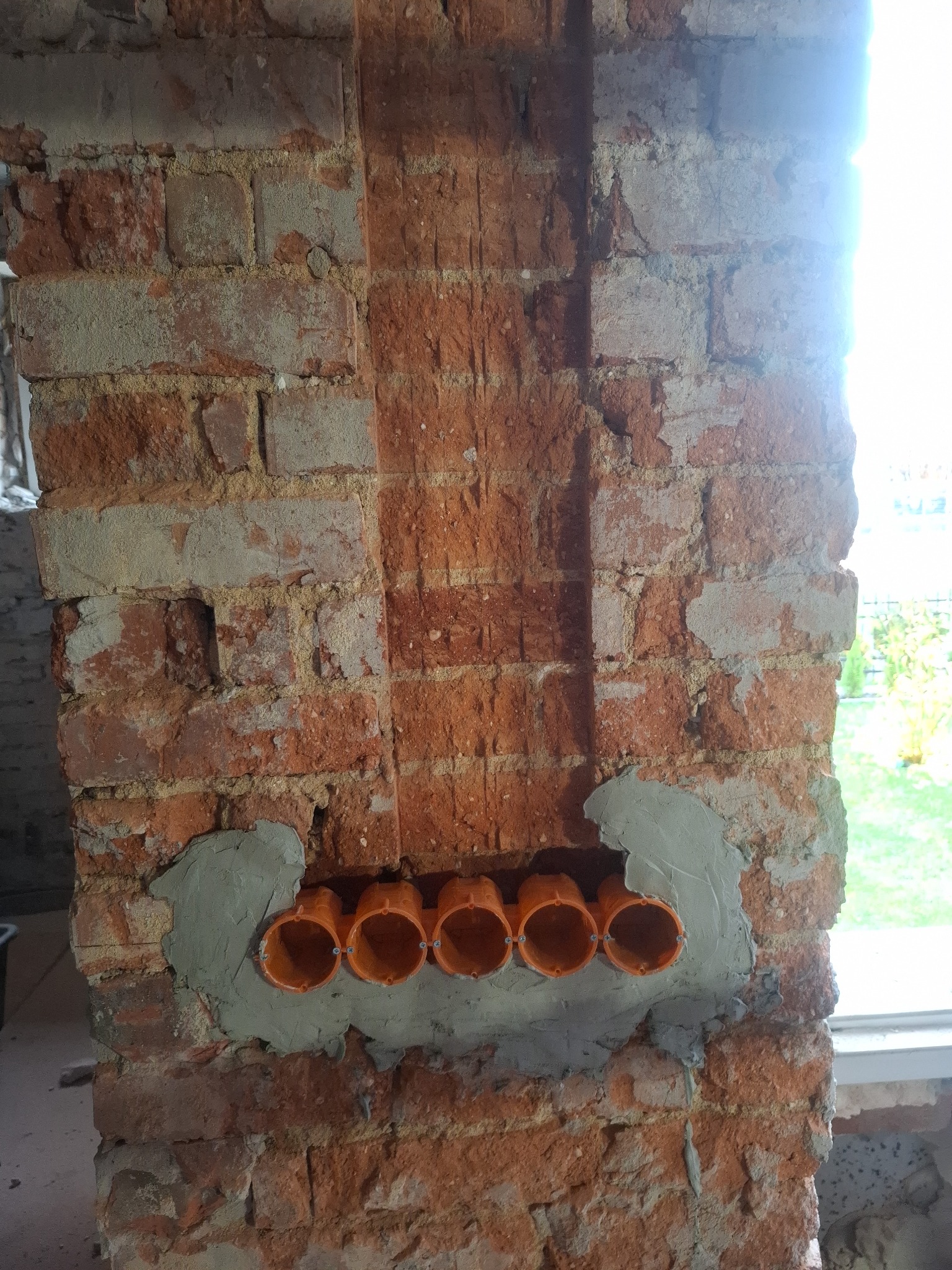 Czerwona cegła z zamontowanymi, pomarańczowymi puszkami elektrycznymi, osadzonymi w szarym cemencie. Widoczne przewody i fragment okna w tle.