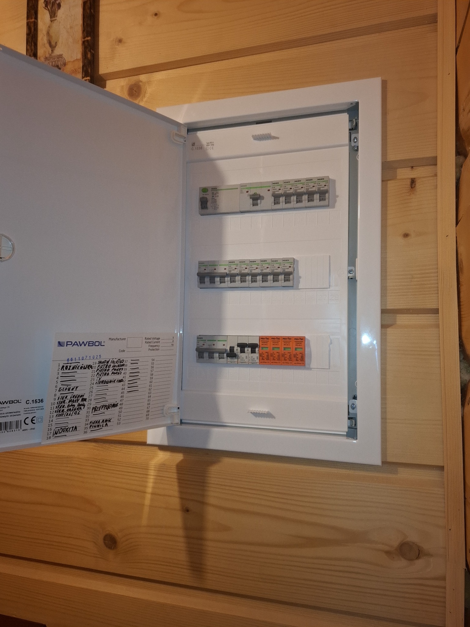 Otwarta skrzynka elektryczna Pawbol z widocznymi bezpiecznikami i opisami obwodów, zamontowana na drewnianej ścianie. Widoczne detale instalacji i oznaczenia.
