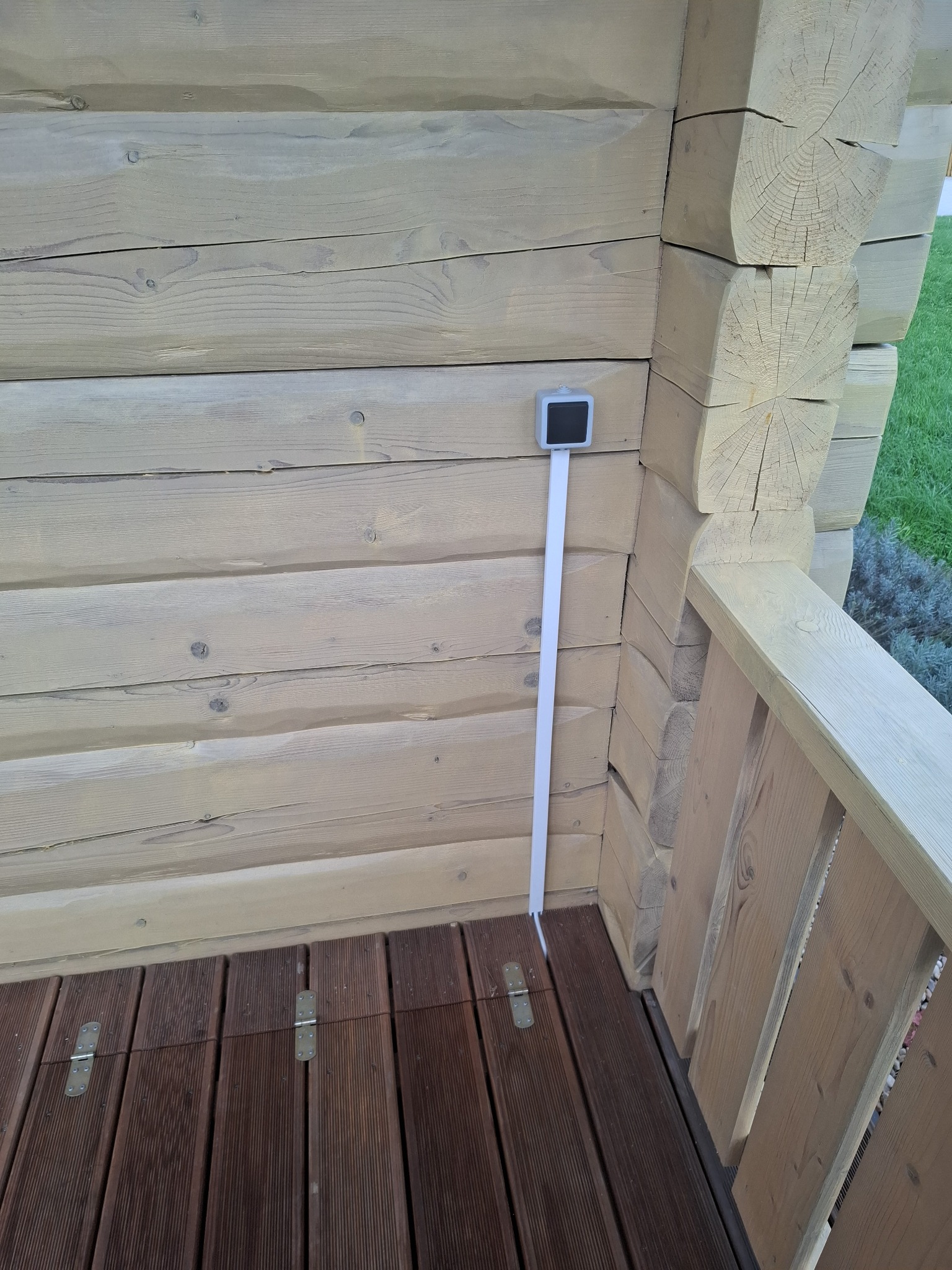Drewniana ściana z szarą puszką elektryczną i kablem w listwie, brązowa podłoga z desek na zewnątrz budynku. Widoczne elementy konstrukcyjne budynku.