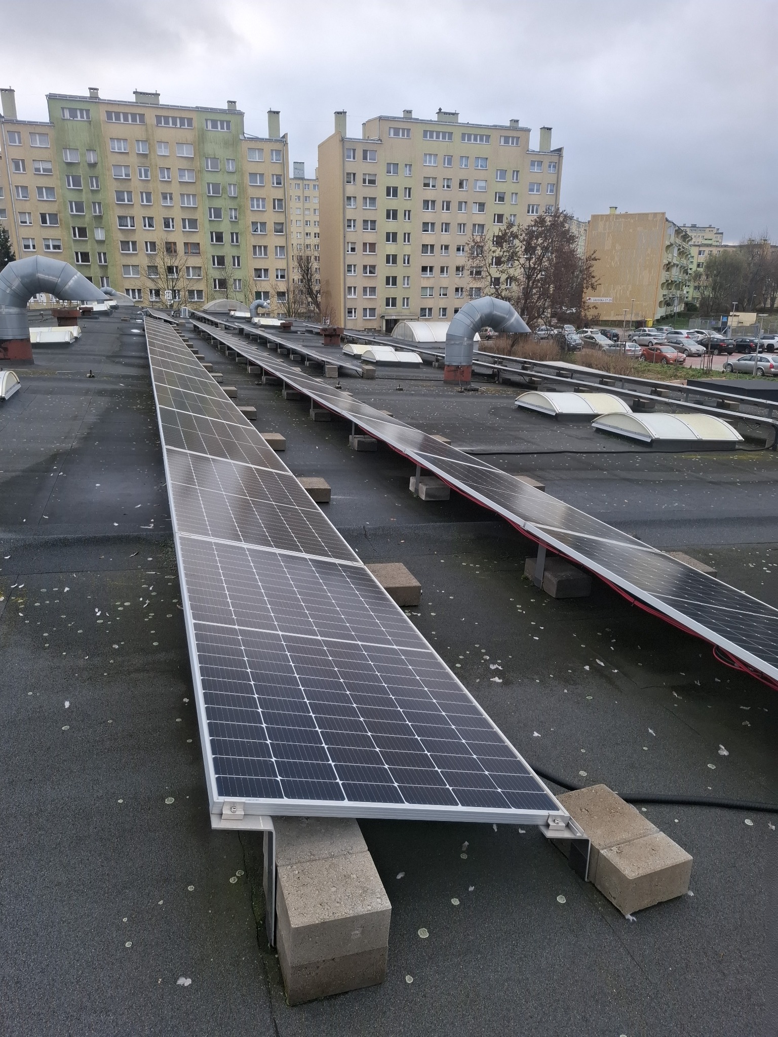 Rząd paneli słonecznych na płaskim dachu budynku mieszkalnego, wspartych na betonowych blokach, z widocznymi elementami wentylacji i parkingiem w tle.