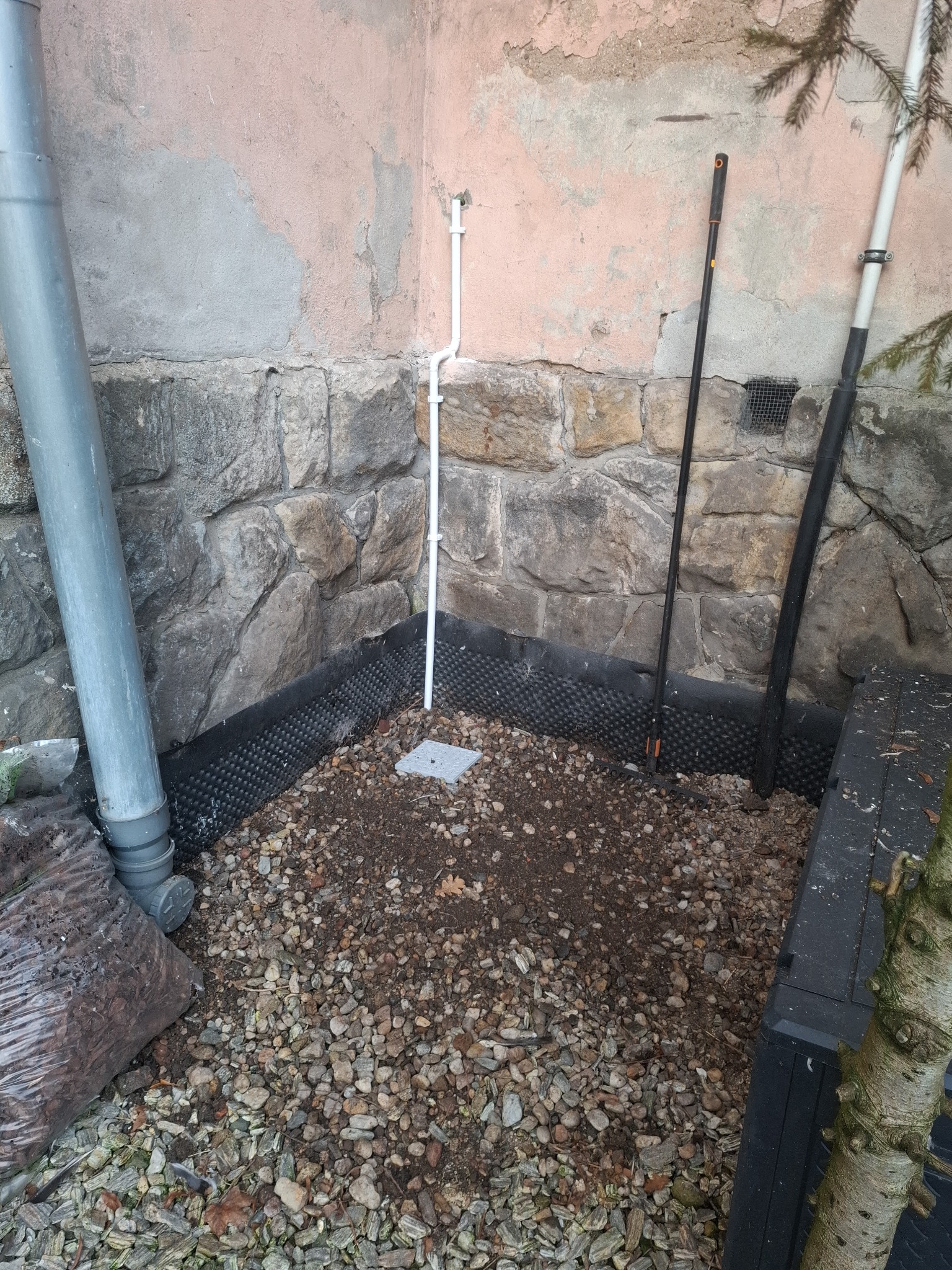 Fragment instalacji drenażowej przy ścianie budynku z kamienną podmurówką. Widoczna rura spustowa, folia kubełkowa i warstwa żwiru. Ujęcie z bliska, z dołu.