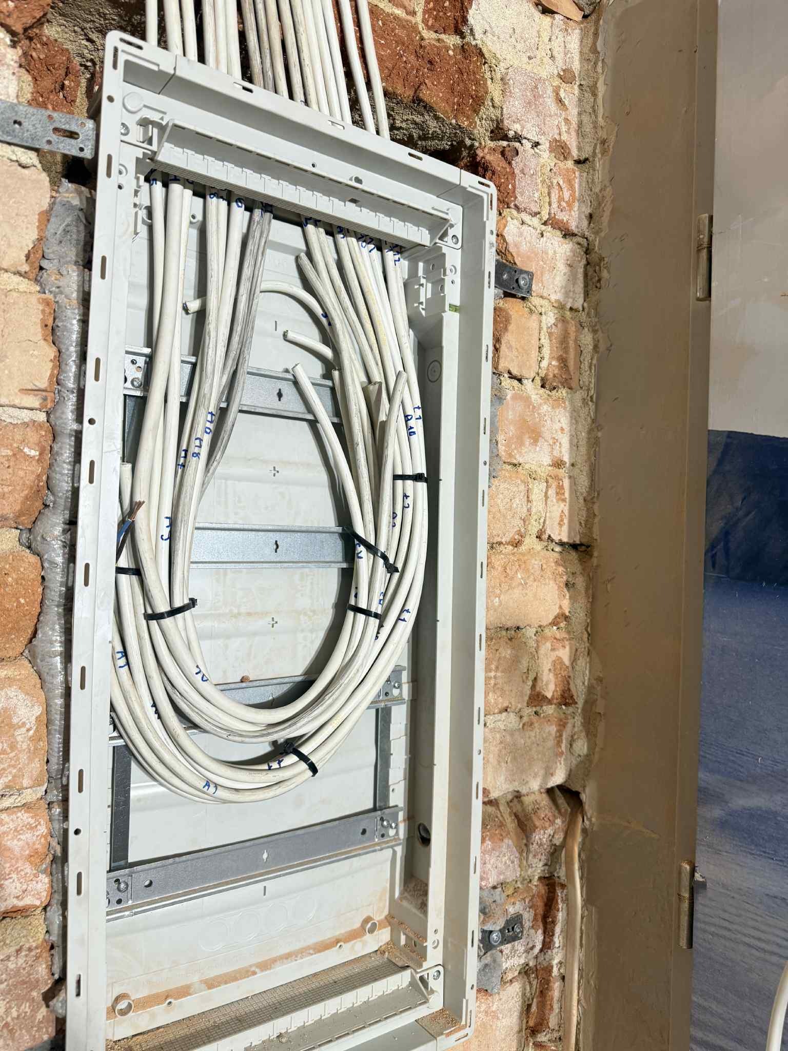 Rozdzielnica elektryczna z ułożonymi przewodami na tle ceglanej ściany. Widoczne metalowe elementy montażowe i fragment ściany z drzwiami. Przewody spięte czarnymi opaskami.