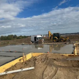 Wylana płyta fundamentowa pod budynek z żółtymi szalunkami, widoczny sprzęt budowlany i bus. Teren przygotowany pod budowę domu, pochmurne niebo.