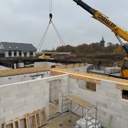 SPSC AB - Budowa domu: ściany z białych bloczków, strop transportowany dźwigiem 'CABALLER', widoczny budynek w tle, stojące rusztowanie i drewniane elementy szalunku.