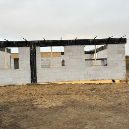 SPSC AB - Wznoszenie ścian domu z betonu komórkowego. Widoczne otwory okienne z drewnianymi podporami. Czarna, pozioma belka na szczycie ściany. Teren budowy w jesiennej scenerii.