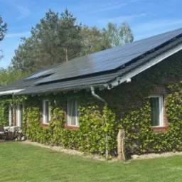 Ceglany dom jednorodzinny z ciemną dachówką, porośnięty bluszczem, z panelami solarnymi na dachu. Na tarasie zestaw mebli ogrodowych. Błękitne niebo.