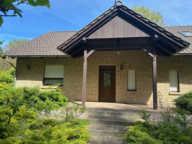 Parterowy dom z brązowym dachem krytym dachówką, z jasnej cegły, z drewnianym gankiem i brązowymi drzwiami wejściowymi. Ogród z bujną zielenią.