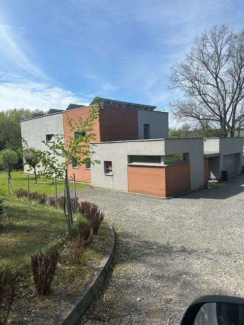 Nowoczesny, dwukondygnacyjny dom z panelami słonecznymi na dachu, wykończony drewnem i szarym tynkiem, otoczony zielenią i żwirowym podjazdem.