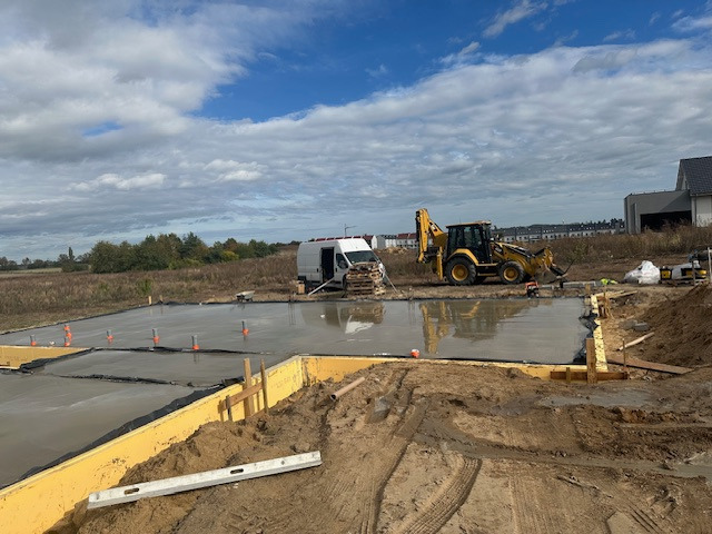 Wylana płyta fundamentowa pod budynek z żółtymi szalunkami, widoczny sprzęt budowlany i bus. Teren przygotowany pod budowę domu, pochmurne niebo.