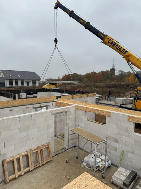 Budowa domu: ściany z białych bloczków, strop transportowany dźwigiem 'CABALLER', widoczny budynek w tle, stojące rusztowanie i drewniane elementy szalunku.