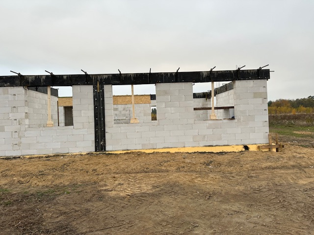 Wznoszenie ścian domu z betonu komórkowego. Widoczne otwory okienne z drewnianymi podporami. Czarna, pozioma belka na szczycie ściany. Teren budowy w jesiennej scenerii.