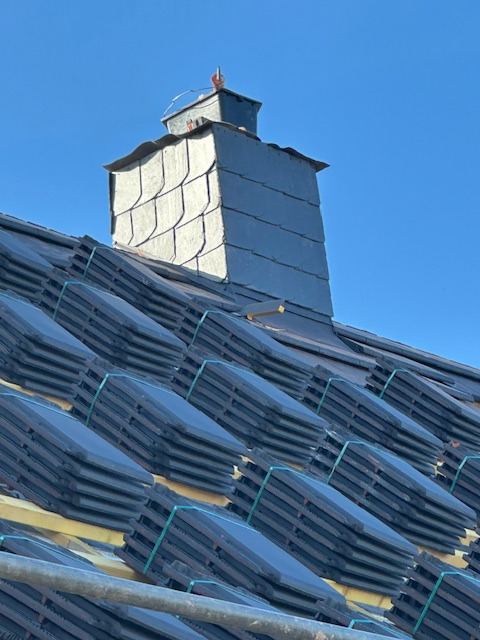 Dach z ułożonymi, czarnymi dachówkami karpiówkami, widoczny komin z łupka. Częściowo ułożone dachówki na łatach. Błękitne niebo w tle.