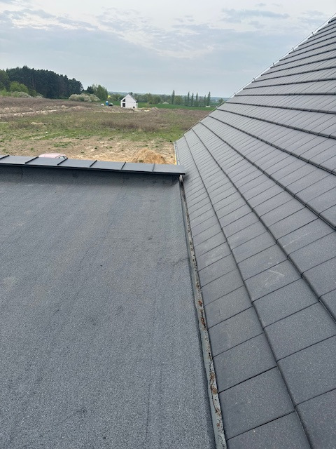 Ciemnoszara dachówka ceramiczna ułożona na dachu domu jednorodzinnego, widok z góry na fragment dachu z papą w tle i krajobrazem polnym w oddali.