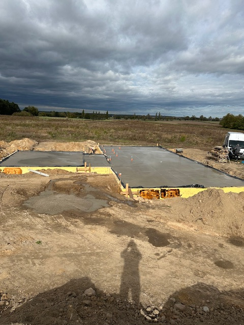 Wylany fundament pod budowę domu jednorodzinnego z widocznym szalunkiem i izolacją, na tle pola i pochmurnego nieba. W tle zaparkowany bus.