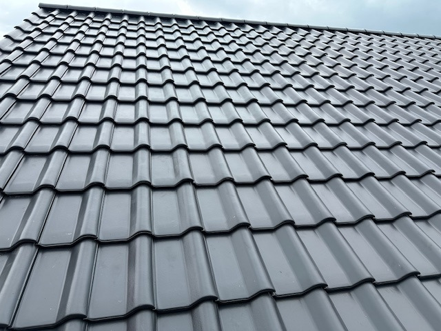 Nowy, grafitowy dach z dachówki ceramicznej, ułożony w regularny wzór. Widok pod kątem, ukazujący strukturę i połysk dachówek. Pochmurne niebo w tle.