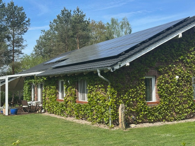 Ceglany dom jednorodzinny z ciemną dachówką, porośnięty bluszczem, z panelami solarnymi na dachu. Na tarasie zestaw mebli ogrodowych. Błękitne niebo.