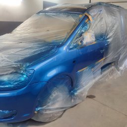 Blacharstwo lakiernictwo samochodowe - Niebieski samochód, częściowo zakryty folią ochronną i zabezpieczony taśmą malarską, przygotowany do lakierowania w warsztacie.