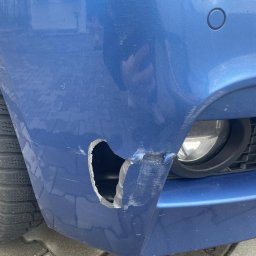 Blacharstwo lakiernictwo samochodowe - Uszkodzony, niebieski przedni zderzak samochodu z widoczną dziurą i zarysowaniami, opona w tle.