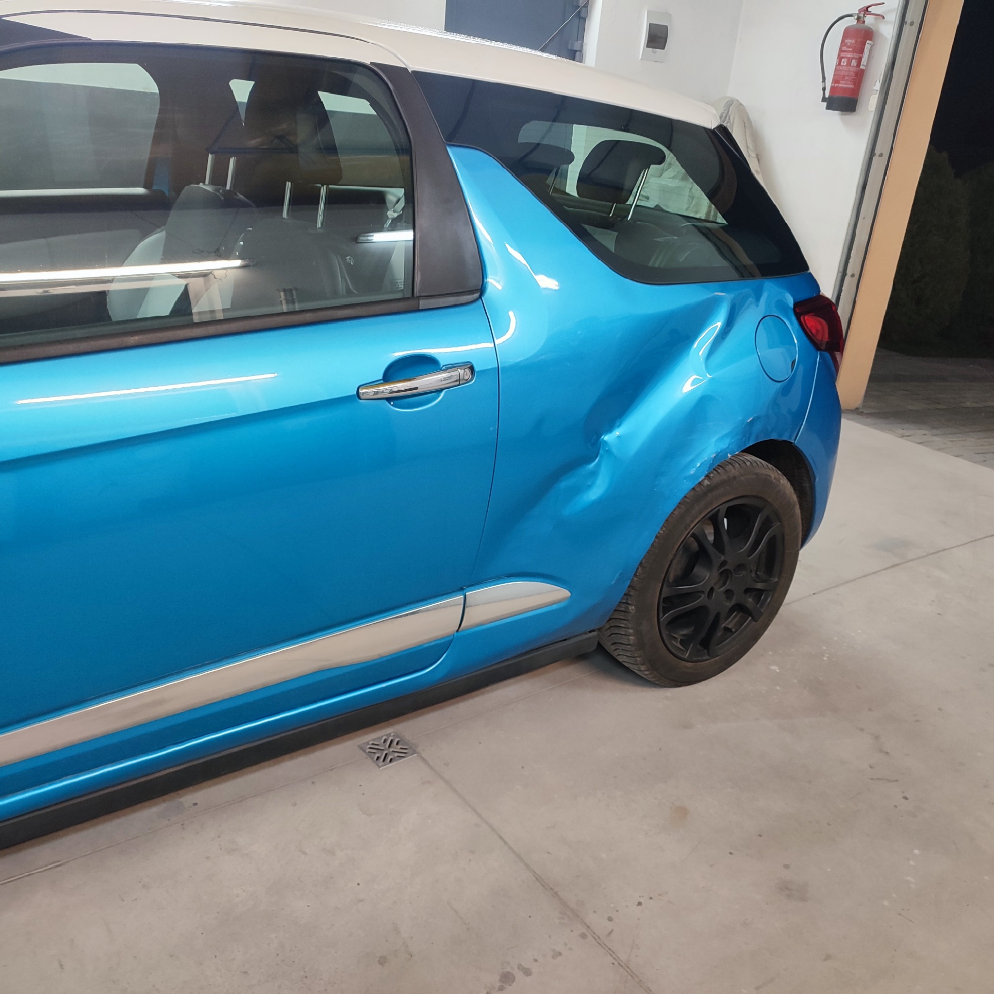 Niebieski samochód z wgnieceniem na tylnym błotniku, widoczne czarne felgi i biały dach, zaparkowany w garażu z gaśnicą na ścianie.