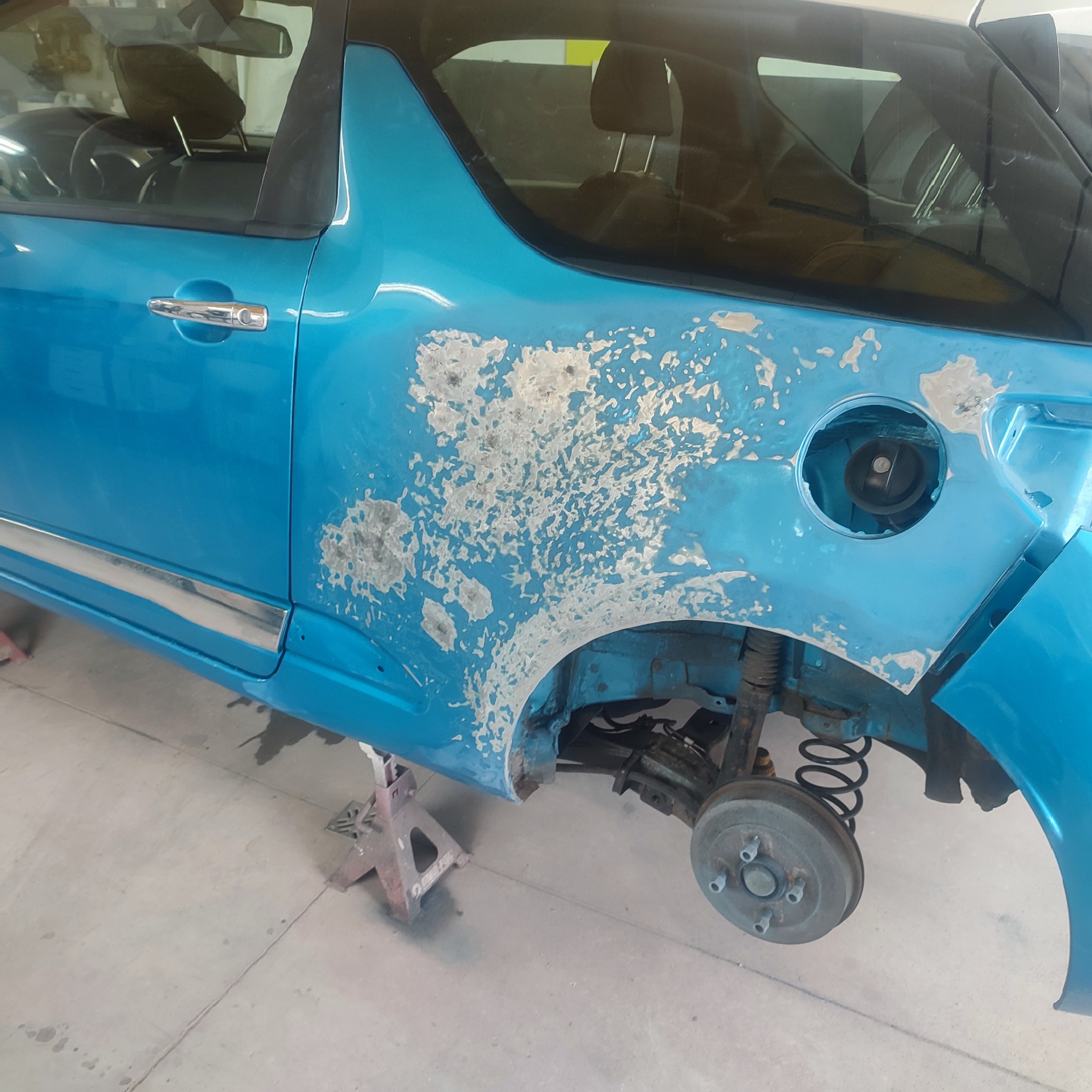 Niebieski samochód na podnośniku z uszkodzoną karoserią, przygotowywaną do naprawy, widoczne ślady szlifowania i brak koła.